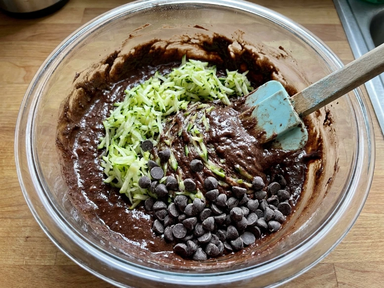 Folding finely shredded raw green zucchini and dark chocolate chips into a thick chocolate brownie batter using a blue rubber spatula in a glass bowl.