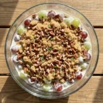 An overhead view of a large glass bowl filled with creamy grape salad, heavily layered with brown sugar, toasted chopped pecans, and fresh mint.