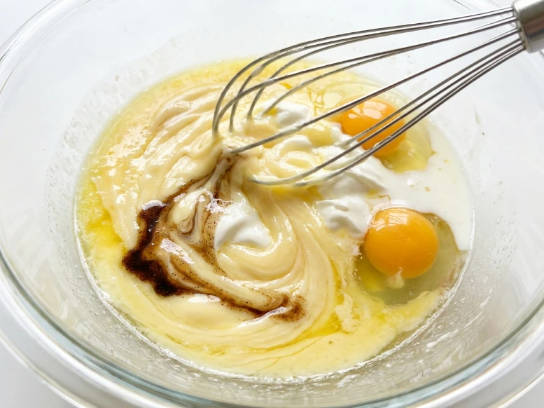 Whisking melted butter, sour cream, eggs, and vanilla extract together in a glass mixing bowl until smooth and uniform.