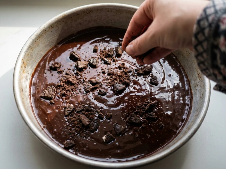 A close-up view of a hand sprinkling crushed Oreo cookies over a rich layer of glossy hot fudge sauce inside a metal pan to create the signature ice cream cake crunch center.
