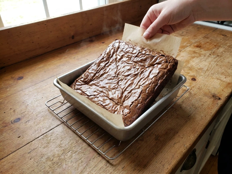 Using parchment paper handles to lift a hot, solid block of freshly baked chocolate brownies out of the metal tin onto a wire cooling rack.