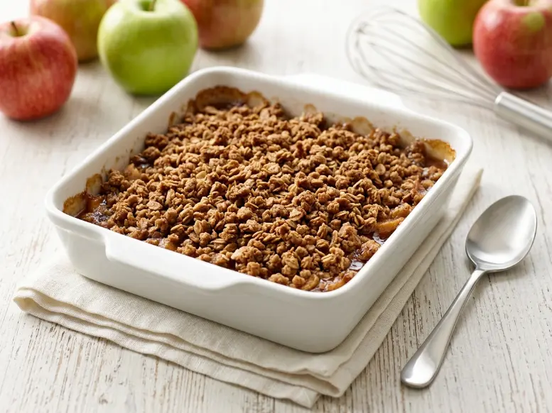 A freshly baked golden delicious apple crisp in a white square ceramic baking dish surrounded by fresh apples and a kitchen whisk.
