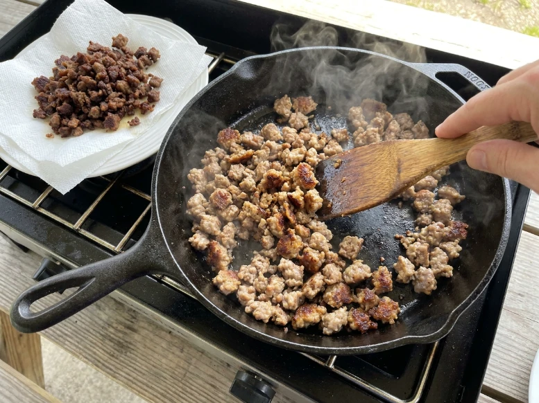 Browning bulk breakfast sausage in a black cast iron skillet on an outdoor stove burner while draining cooked meat on a paper-towel-lined plate.